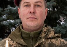 Portrait of a soldier in camouflage and a maroon beret with an emblem, standing in front of trees; memorial banner reads 'ВІЧНА ПАМ’ЯТЬ ГЕРОЮ!' with a lit candle in the foreground.