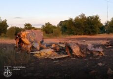 Wrecked, burnt-out vehicle scattered on a dirt field at sunset, with trees in the background and the Ukrainian Prosecutor’s Office emblem in the lower-left corner.