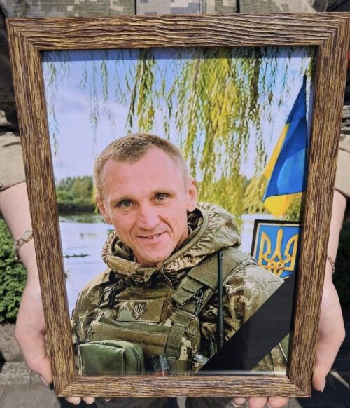 Portrait of a soldier in camouflage gear inside a wooden frame, with a Ukrainian flag visible in the background.