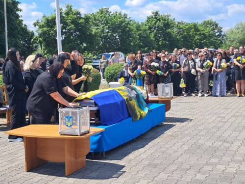 M mourners gather for an outdoor funeral, with a casket draped in blue and yellow Ukraine colors on a blue table and flowers nearby.