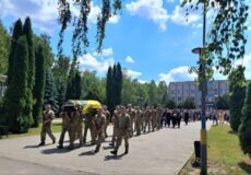 Line of soldiers in uniform marching in a military funeral, with a coffin draped in yellow and blue carried at the front on a sunny day.