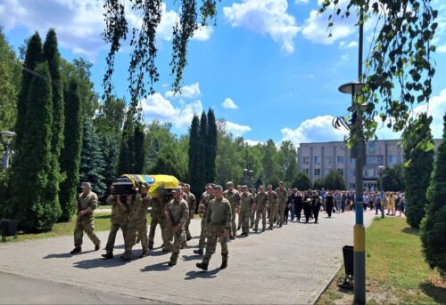 Line of soldiers in uniform marching in a military funeral, with a coffin draped in yellow and blue carried at the front on a sunny day.