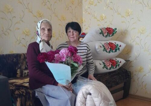 Two women sit on a living room couch; the elderly woman holds a bouquet of pink flowers, another woman sits beside her, with a rolled quilt or mat in front of them.
