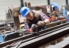 Metalworker wearing a blue hard hat and plaid shirt shaping a long metal beam with a hand tool in a cluttered workshop