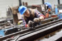 Metalworker wearing a blue hard hat and plaid shirt shaping a long metal beam with a hand tool in a cluttered workshop