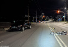 Nighttime city street; a dark SUV on the left and a person covered with a reflective blanket lying in the road ahead.