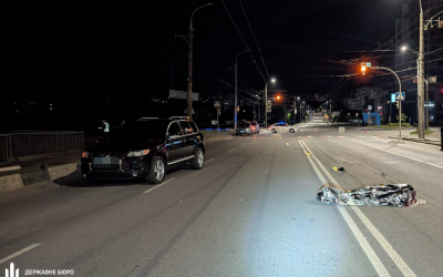 Nighttime city street; a dark SUV on the left and a person covered with a reflective blanket lying in the road ahead.