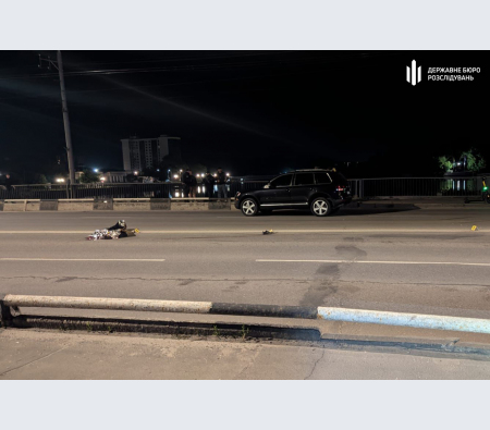Nighttime road incident: a dark SUV on a multi-lane street with debris scattered along the road; guardrail in the foreground and responders in the distance.