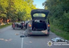 SUV with rear hatch open on a rural road amid debris and a wrecked motorcycle trailer in the background; Ukrainian police emblem visible.