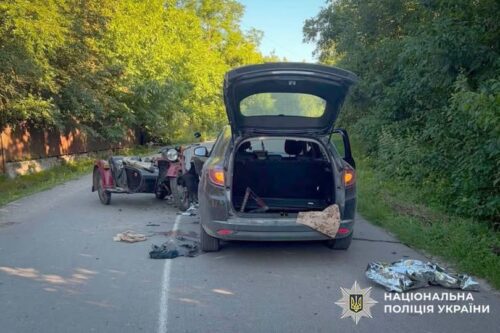 SUV with rear hatch open on a rural road amid debris and a wrecked motorcycle trailer in the background; Ukrainian police emblem visible.