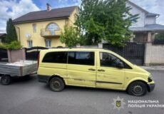 Yellow passenger van with side damage and a trailer attached, parked on a residential street with houses in the background; police watermark visible in corner.