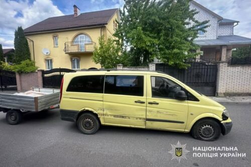 Yellow passenger van with side damage and a trailer attached, parked on a residential street with houses in the background; police watermark visible in corner.