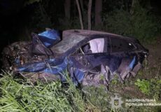 Night scene of a severely damaged blue car off a grassy roadside, crumpled in a ditch with open windows; police watermark visible in corner, Ukraine.