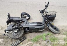 Black electric scooter tipped over on a curb, front wheel damaged; Ukrainian police watermark.