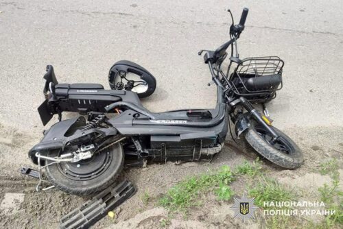 Black electric scooter tipped over on a curb, front wheel damaged; Ukrainian police watermark.
