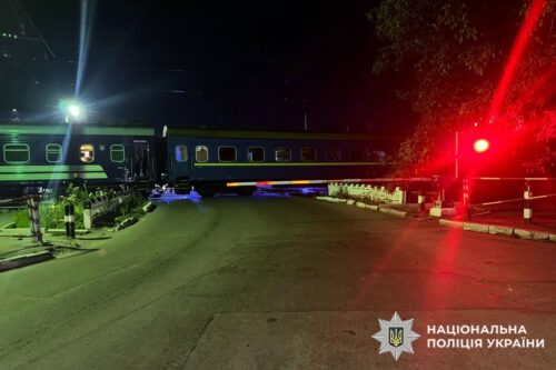 Night scene of a green and blue passenger train stopped at a level crossing with red signal lights and barriers down; Ukrainian police logo visible.