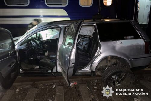 Silver SUV with open doors and visible damage beside a blue train car at night; Ukrainian police watermark