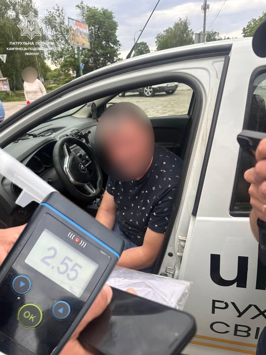 Man seated in the driver’s seat of a white police car while a breathalyzer displays 2.55 on its screen (face blurred).