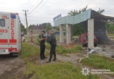 Two police officers inspect a damaged site beside an ambulance, with rubble and a collapsed shelter and a red car in the background.