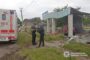 Two police officers inspect a damaged site beside an ambulance, with rubble and a collapsed shelter and a red car in the background.