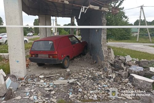 Red hatchback car crashed into a damaged wall of a building with rubble and debris scattered on the ground; police emblem visible in the lower right corner.