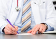 Doctor in a white coat signs a document with a purple pen; stethoscope hangs around the neck.