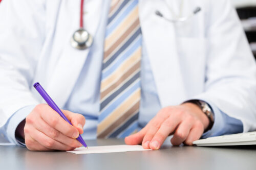Doctor in a white coat signs a document with a purple pen; stethoscope hangs around the neck.