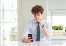 Young man in a light blue shirt and gray tie sits at a white table, gripping a smartphone and frowning in frustration at his phone.