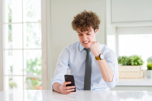 Young man in a light blue shirt and gray tie sits at a white table, gripping a smartphone and frowning in frustration at his phone.