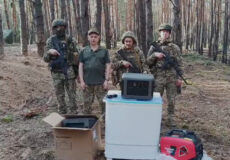 Four armed soldiers stand in a pine forest beside a washing machine and cardboard boxes, posing for the camera.