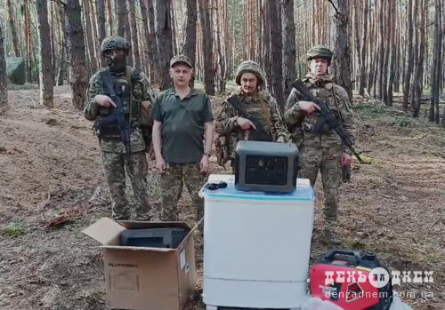 Four armed soldiers stand in a pine forest beside a washing machine and cardboard boxes, posing for the camera.