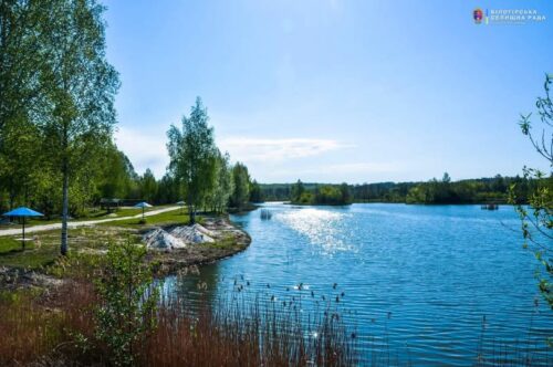 Sunlit lake with a grassy shoreline, trees, and blue umbrellas along a path.