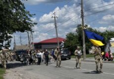 Soldiers march down a sunny street carrying a large Ukrainian flag, with civilians watching along a residential road.