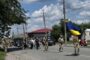 Soldiers march down a sunny street carrying a large Ukrainian flag, with civilians watching along a residential road.