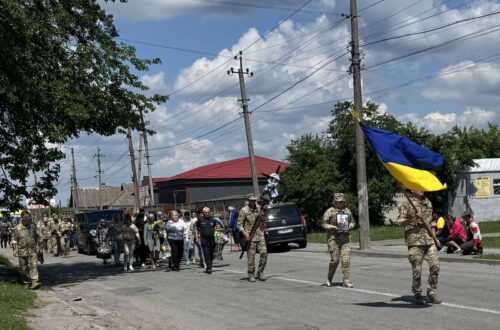 Soldiers march down a sunny street carrying a large Ukrainian flag, with civilians watching along a residential road.