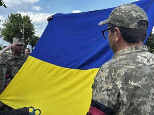 Two soldiers in camouflage hold a large blue-and-yellow Ukrainian flag outdoors during daytime.