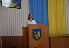 Woman delivering a speech at a wooden podium bearing Ukraine's coat of arms, with blue and yellow banners behind her.