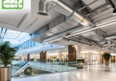 Bright shopping mall atrium with an escalator, glass railing, storefronts, potted plants, and exposed silver HVAC ducts overhead