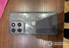 Top-down view of a gray Motorola phone with a quad-camera module on a wooden table; police watermark in bottom-right corner.