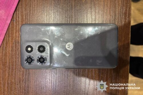 Top-down view of a gray Motorola phone with a quad-camera module on a wooden table; police watermark in bottom-right corner.