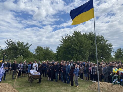 Crowd at an outdoor funeral graveside, a coffin on a bier, with a blue-and-yellow Ukrainian flag nearby.