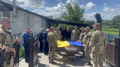 Military funeral outdoors with a coffin draped in blue-and-yellow flag colors, surrounded by uniformed soldiers and civilians.