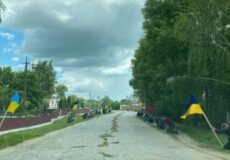 Long rural road with people kneeling along both sides, Ukrainian flags and flowers laid along the center under a cloudy sky.