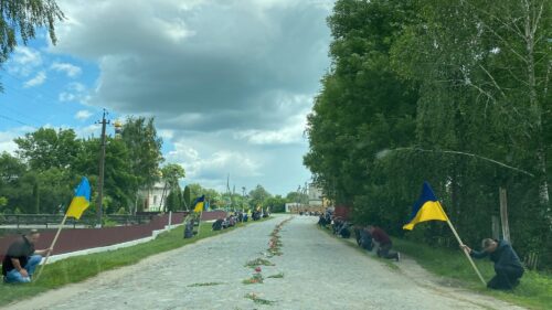 Long rural road with people kneeling along both sides, Ukrainian flags and flowers laid along the center under a cloudy sky.