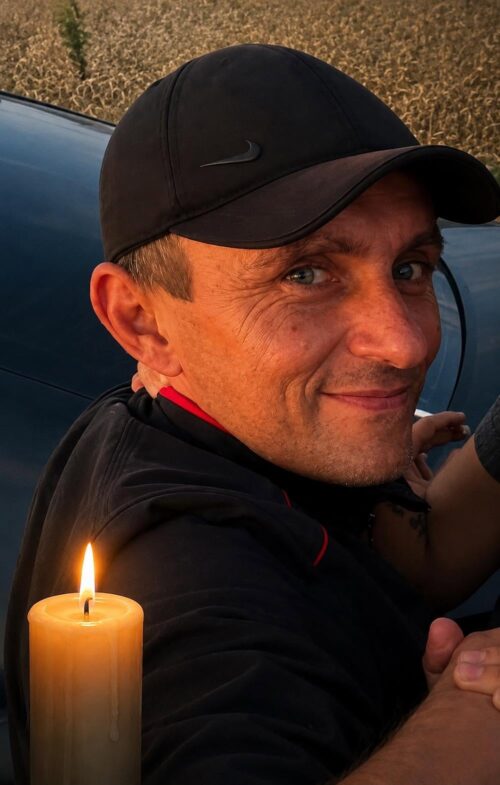Smiling man in a black cap beside a lit candle outdoors.