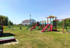 Colorful children's playground with slides, swings, and climber on a grassy field under a clear blue sky; a building in the background.