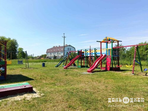 Colorful children's playground with slides, swings, and climber on a grassy field under a clear blue sky; a building in the background.