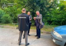 Group of people talking beside a parked blue car; a man with a jacket labeled 'ПРОКУРАТУРА' stands with his back to the camera, others hold documents, green trees in background (Ukraine)