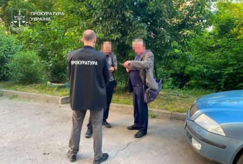 Group of people talking beside a parked blue car; a man with a jacket labeled 'ПРОКУРАТУРА' stands with his back to the camera, others hold documents, green trees in background (Ukraine)