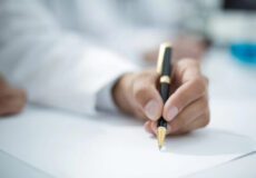 Close-up of a hand in a white coat writing with a gold-and-black fountain pen on paper.